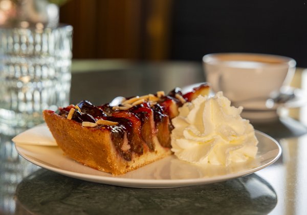 Ein Stück Pflaumenkuchen, garniert mit Mandelstückchen und einer Portion Schlagsahne, dazu eine Tasse Kaffee im Hintergrund.